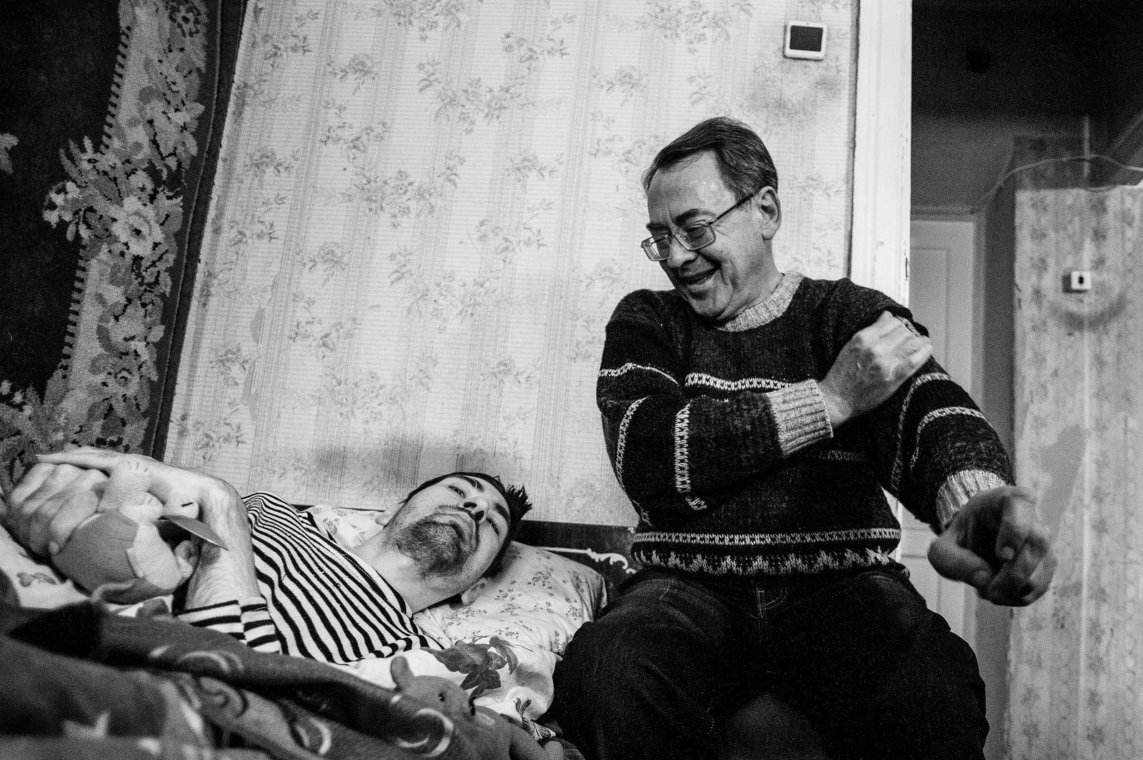 Arik Shraga. "Joint" Families: The Stories of Love. Kherson. Dima with his father Dima was absolutely healthy until the age of 3 when a routine immunization at a kindergarten has brought an infection which made Dima disabled. Epilepsy developed, with severe leg pains and seizures. His condition kept worsening all the time. By the age of 30 he could not even sit up on his bed, he spent his days lying and listening to the radio.
He became so weak that he couldn’t even speak, short of answering “yes” or “no” when his father addresses to him.
His mother died of cancer when he was 30, and only his father remained to take care of him. The father was only showing his true love and admiration to Dima, hiding away his tears, fear and depression. He is visually impaired himself, with his own health deteriorating with years and constant stress. He was afraid most of passing away, which would leave Dima all alone. This didn’t happen. Dima was first to pass away, at the age of 32.